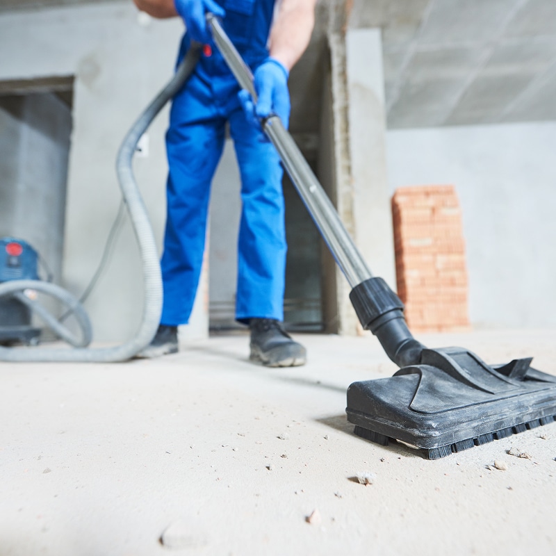 Homme en bleu avec gants aspire un sol poussiéreux de chantier. Nettoyage professionnel après travaux de construction.