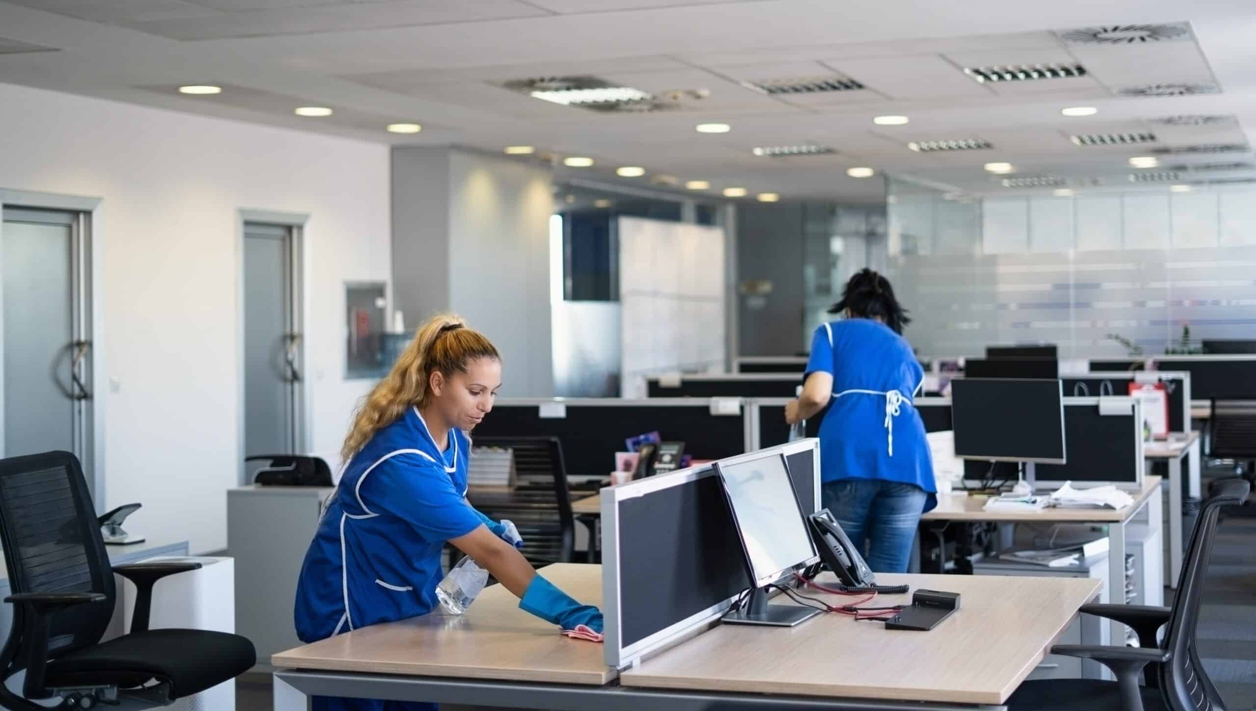 Deux agents d'entretien en uniforme bleu nettoient un bureau moderne. Une femme essuie un poste de travail avec spray et chiffon, l'autre en arrière-plan.