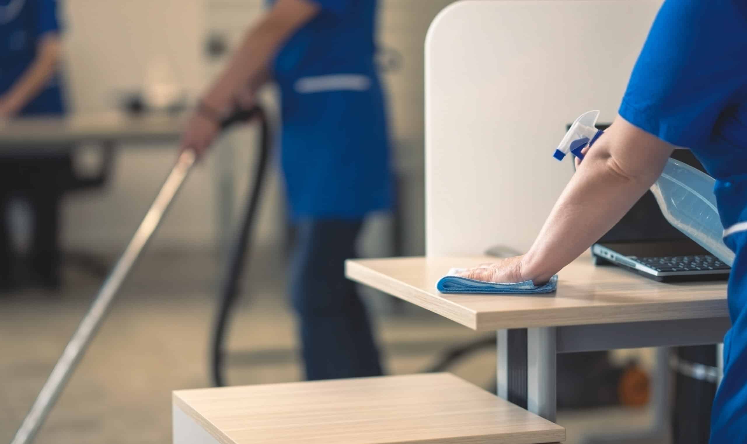 Une personne nettoie un bureau avec un chiffon bleu et un vaporisateur, tandis qu'une autre passe l'aspirateur en arrière-plan. Nettoyage de bureau professionnel.