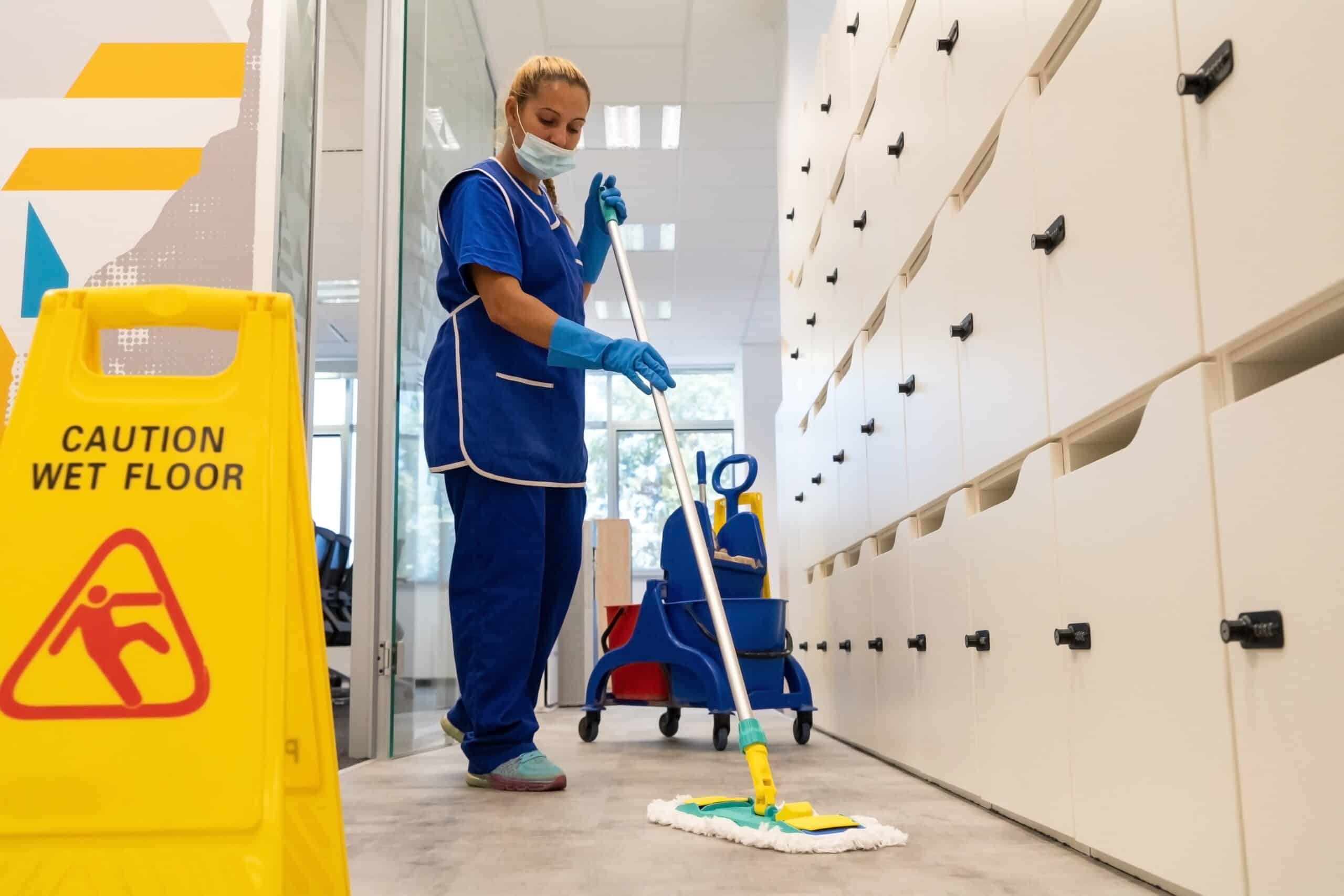 Femme nettoyeuse en uniforme bleu et masque, moppant un sol près d'un panneau jaune "CAUTION WET FLOOR". Seau et casiers clairs en arrière-plan.